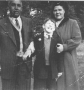 A black and white photo of a young Edward. He is standing, smiling with his mum and dad.