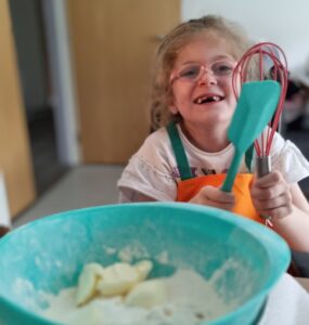 Imogen is smiling and holding a whisk. She is baking in the kitchen.