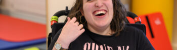 Photo of a young adult in her mid-twenties. She is smiling at the camera. She is a wheelchair user, and has cerebral palsy.